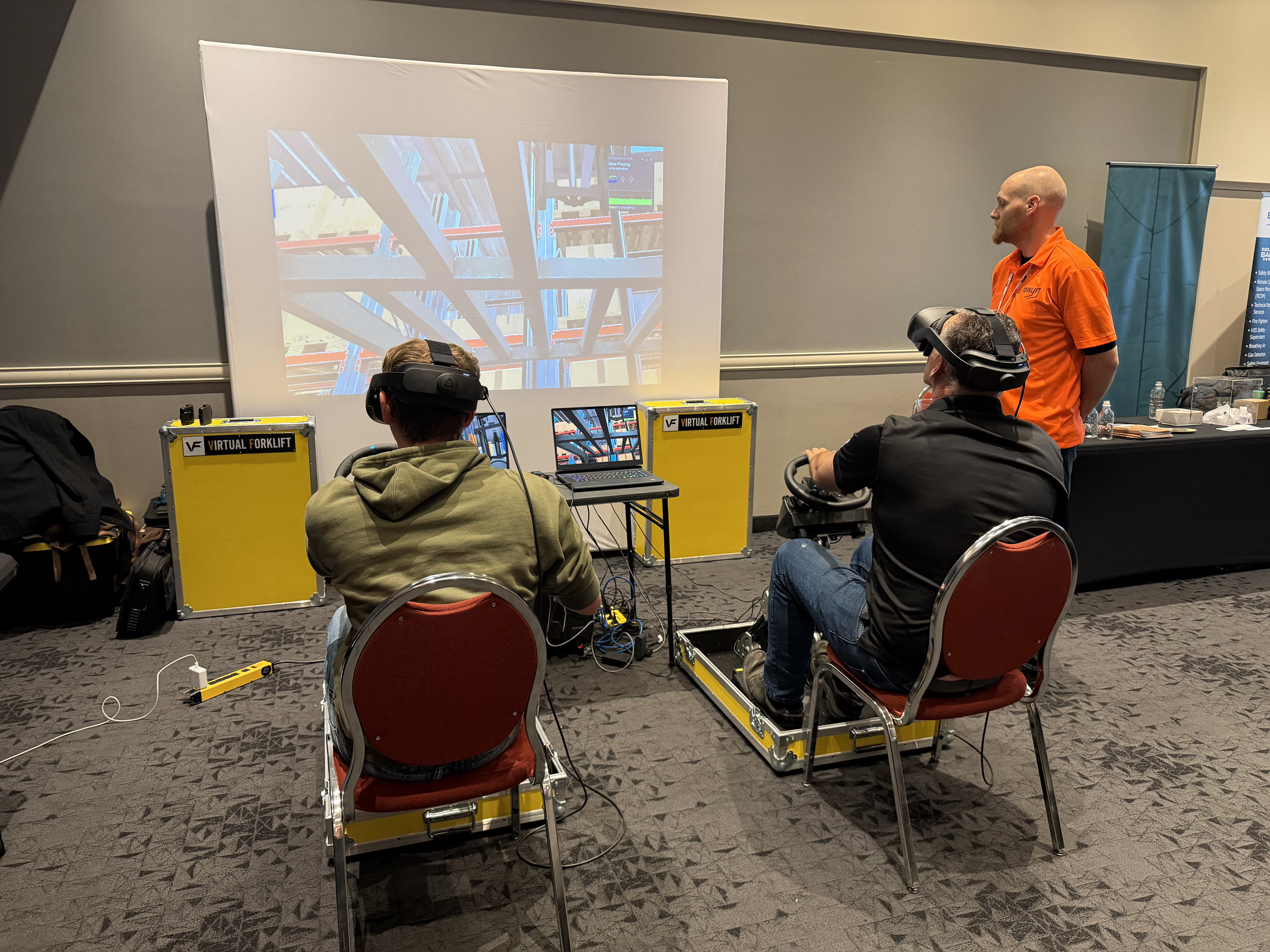 Two trainees in VR headsets on Fold N Go stations at a trade show, with one trainee's headset view on a projector